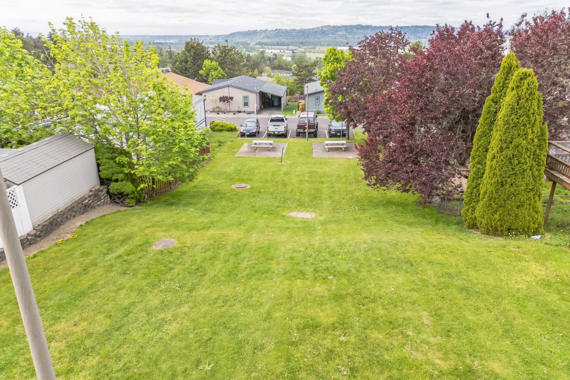 Grassy yard area with picnic tables, bordered by trees and small sheds, with houses and a parking area in the background in Troutdale, OR