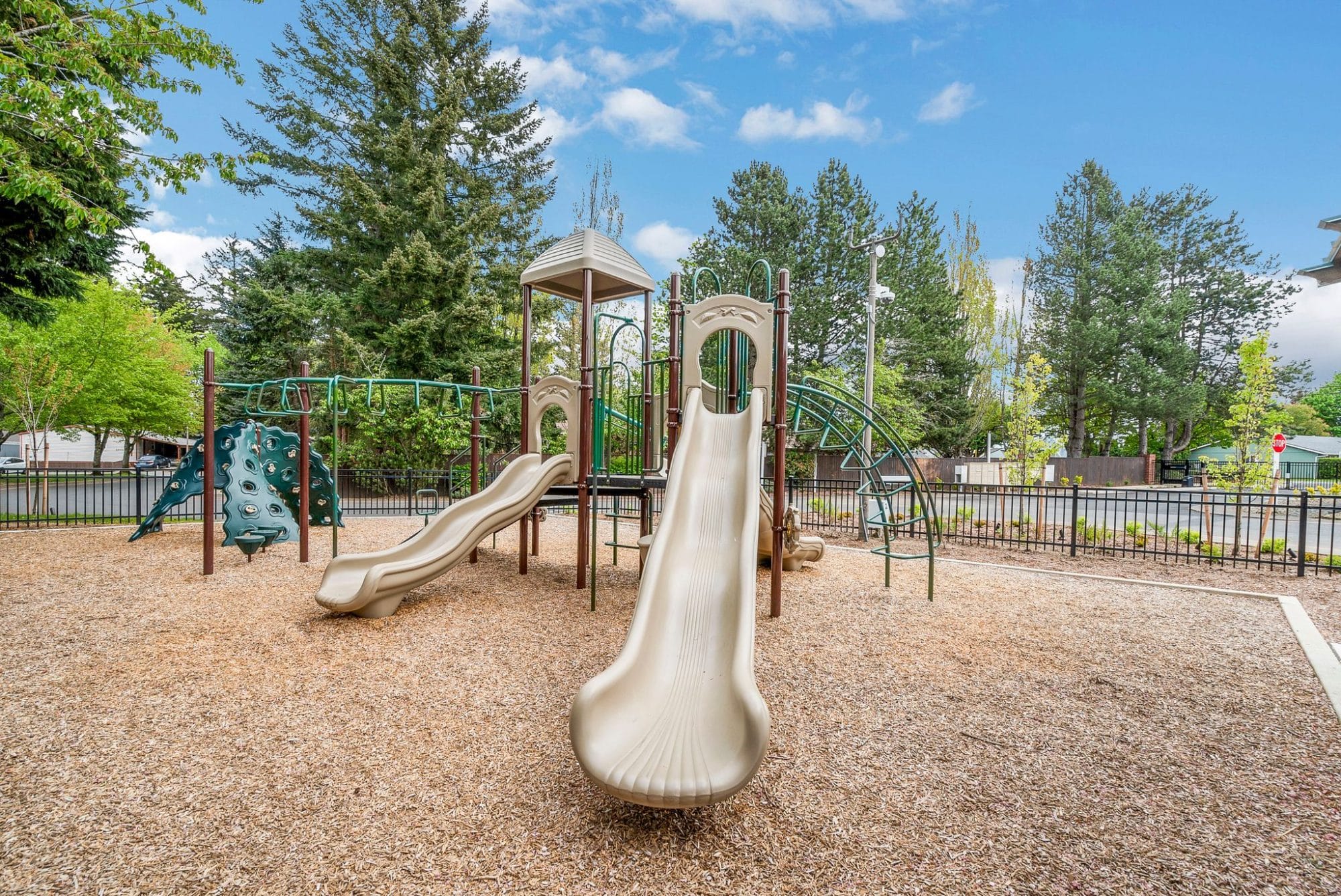 Playground with slides, climbing structures, and wood chip ground covering surrounded by tall trees and enclosed by a fence in Troutdale, OR